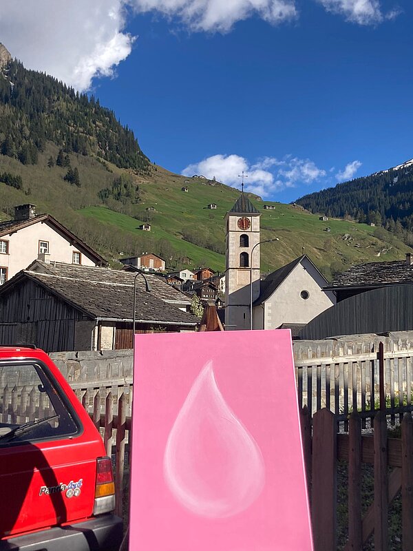 Ein pink gemalter Wassertropfen vor einem blauen Himmel und der Dorfkirche in Vals